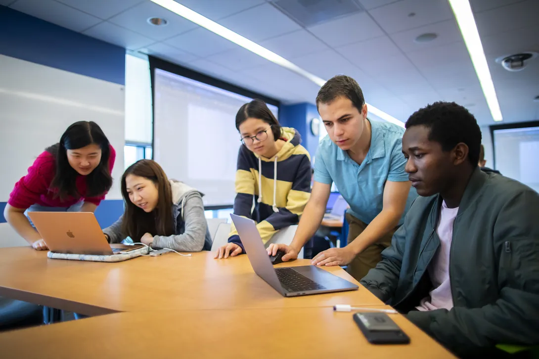 Five students sitting at a table in front of two laptops