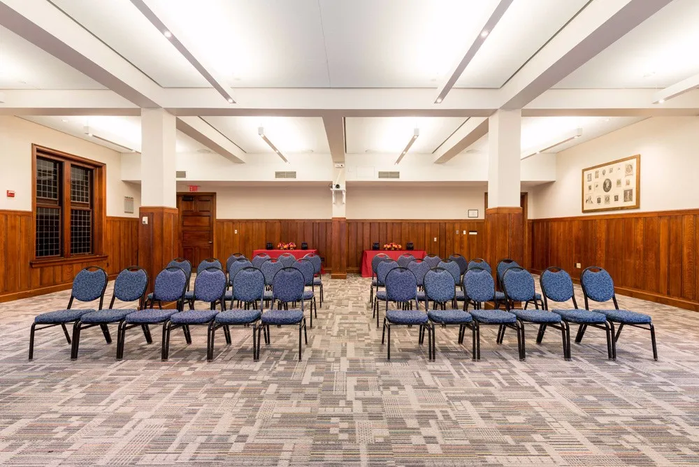 Meeting room with wood wall paneling. Two tables with red table clothes in the back with blue chairs in theater style seating with an aisle between them.