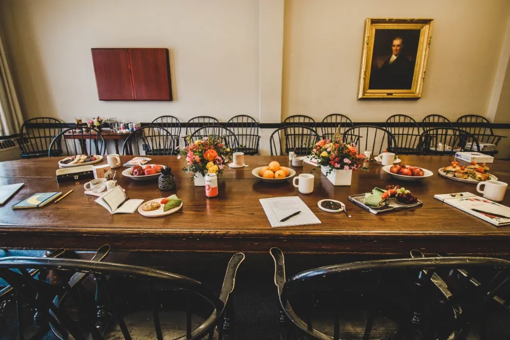 Dark wood board room table with food and documents on it surrounded by chairs