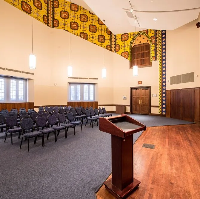 View of room with yellow patterned walls and blue chairs set in theater style in front of a podium