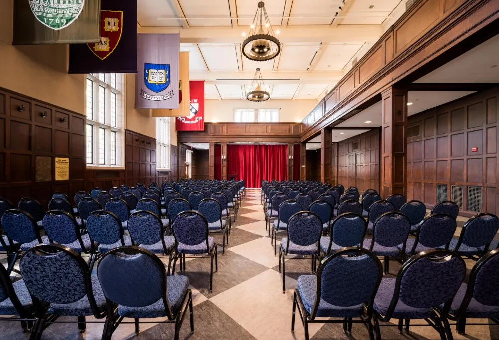 Hall of Flags view from the ground floor showing many blue chair set theatre style against a backdrop of large stained glass windows and Ivy League school flags hanging from the ceiling.