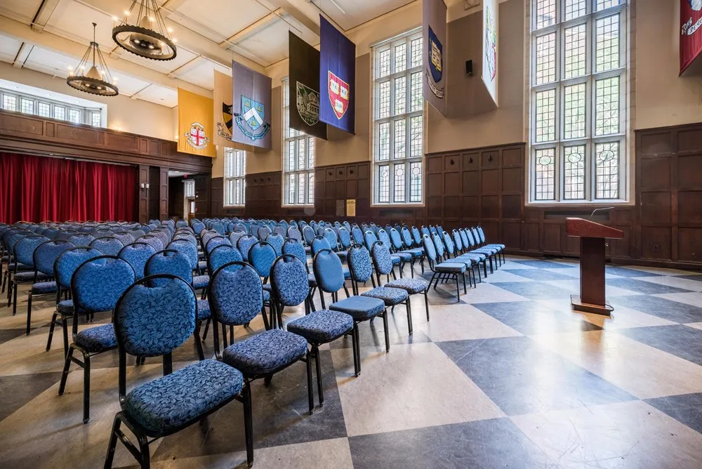 Hall of Flags view from the ground floor showing many blue chair set theatre style against a backdrop of large stained glass windows and Ivy League school flags hanging from the ceiling.